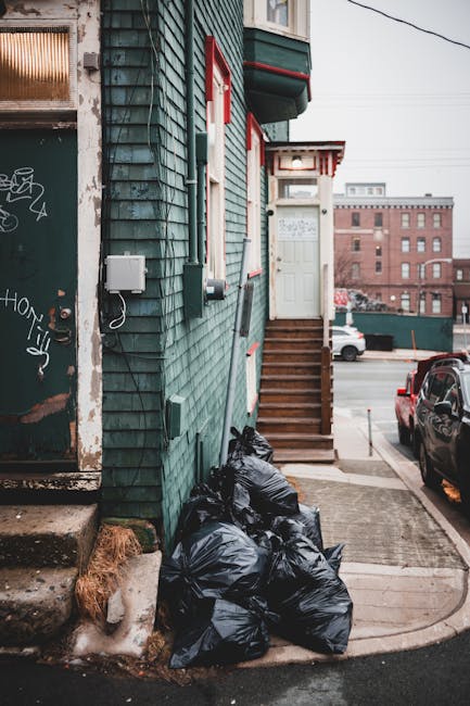 A large pile of mixed household waste and rubbish bags is accumulated on a paved street in front of a row of shops and a parked silver car. The waste includes various black and clear plastic bags, fla