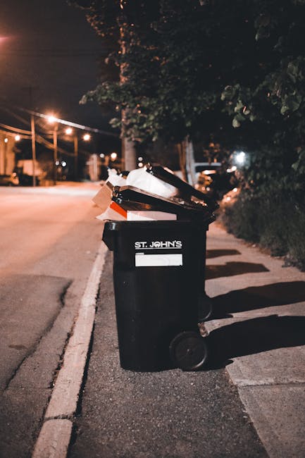 A waste management worker wearing a high-visibility yellow vest and dark clothing is operating a modern rubbish collection vehicle on an urban street during dusk. The worker stands with their back to
